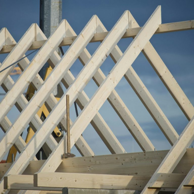 Neubau EH 55: Ein halb fertiger Dachstuhl, im Hintergund ein Kran, ein großer Hammer auf einem Balken symbolisiert, dass gebaut wird